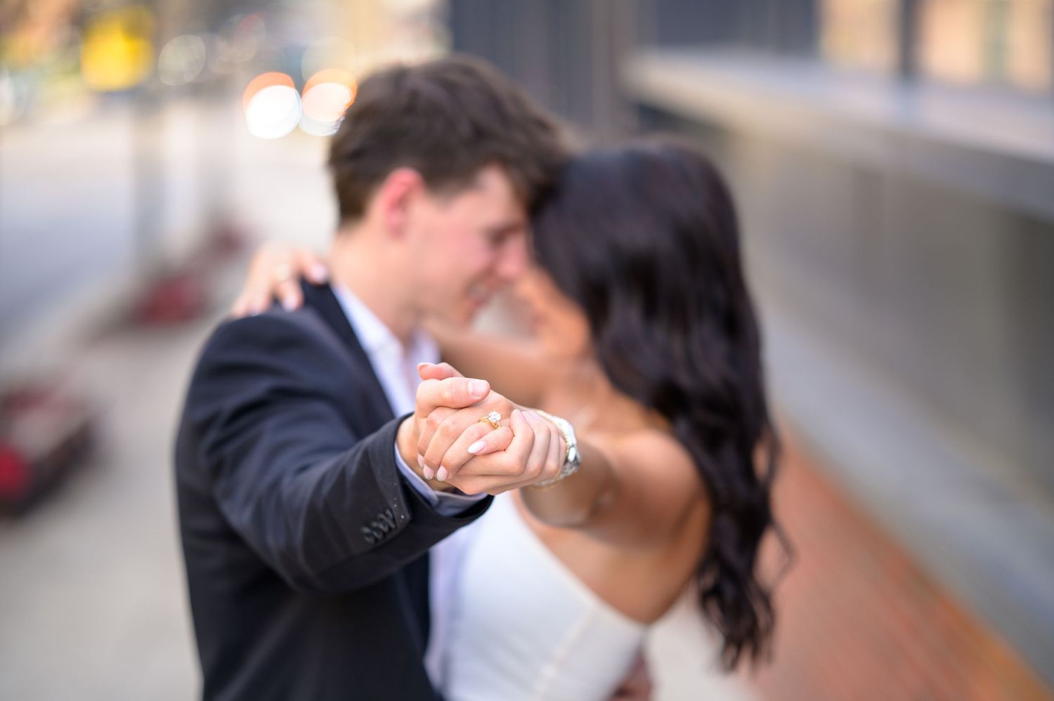Couple sharing a romantic dance outdoors, softly blurred with focus on their clasped hands and engagement ring.