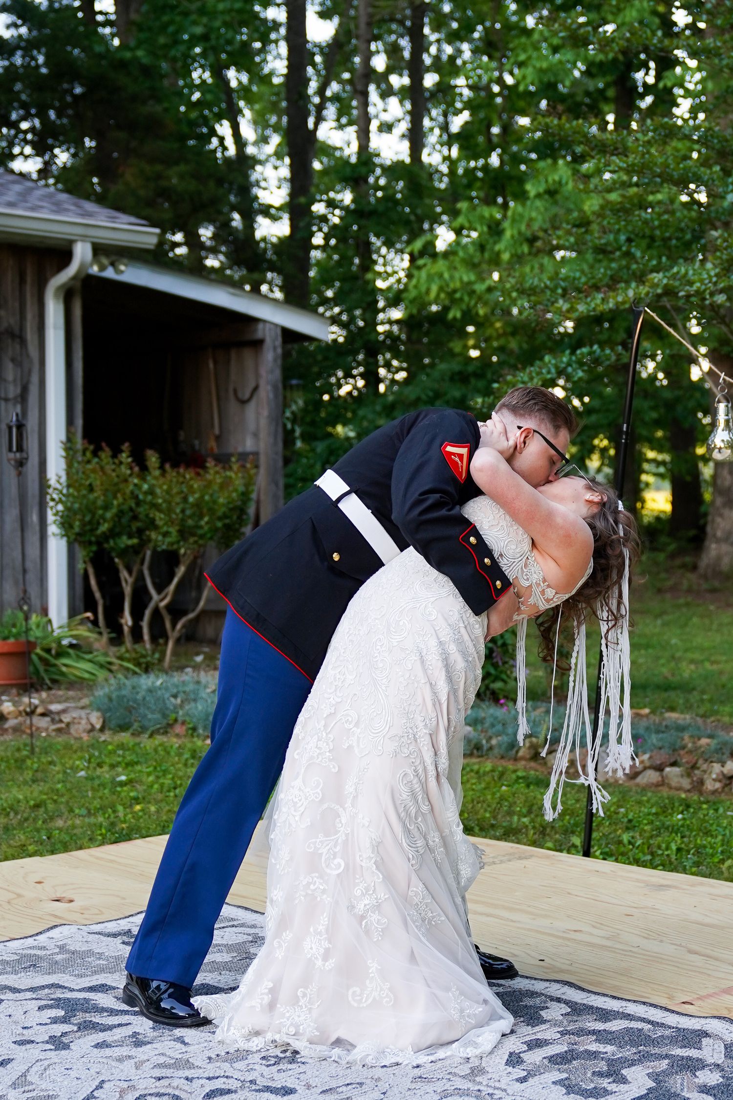 Marine groom dips bride in white lace dress for romantic outdoor wedding kiss on wooden dance floor.