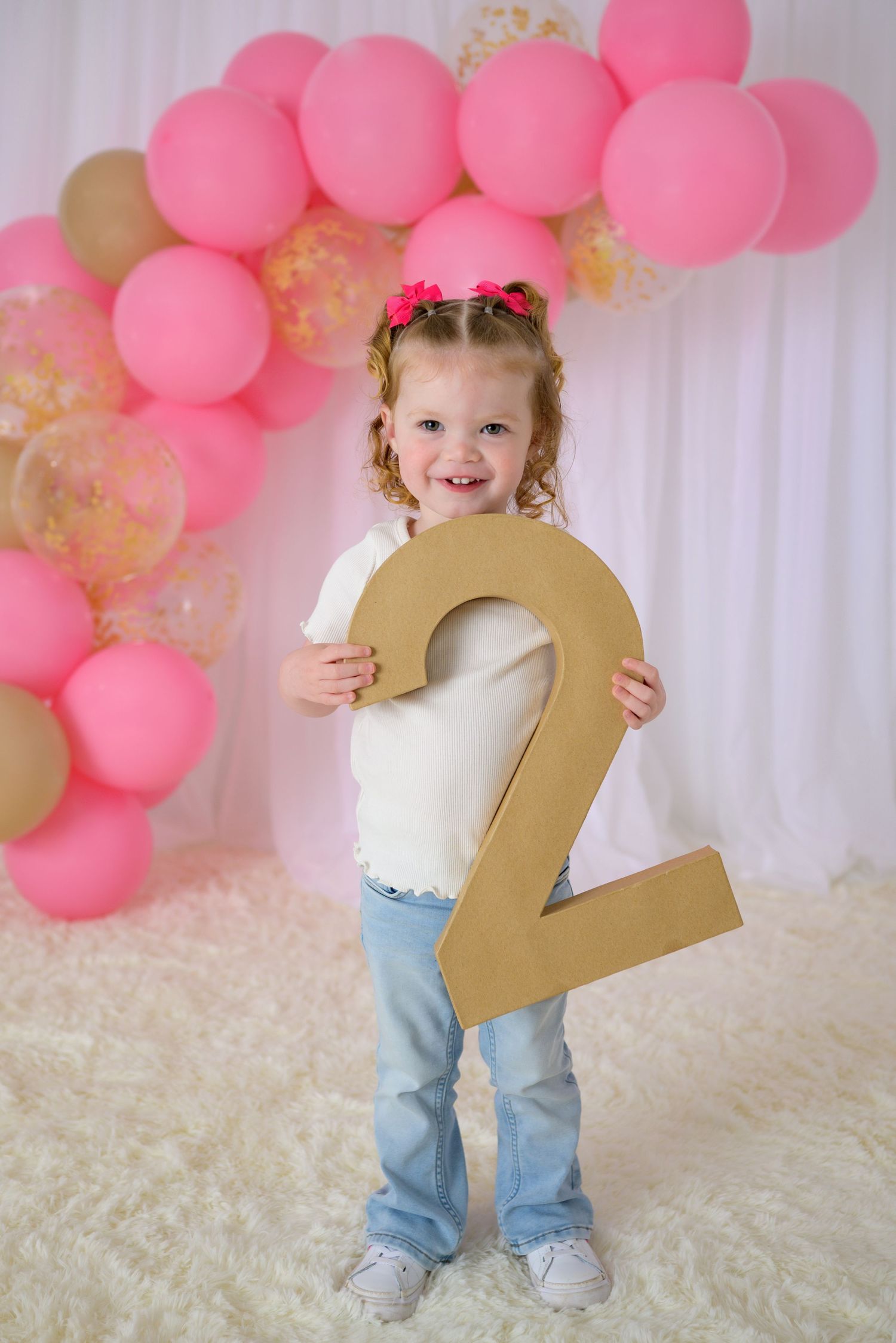 Smiling toddler girl holding large number 2 at pink balloon birthday party celebration.