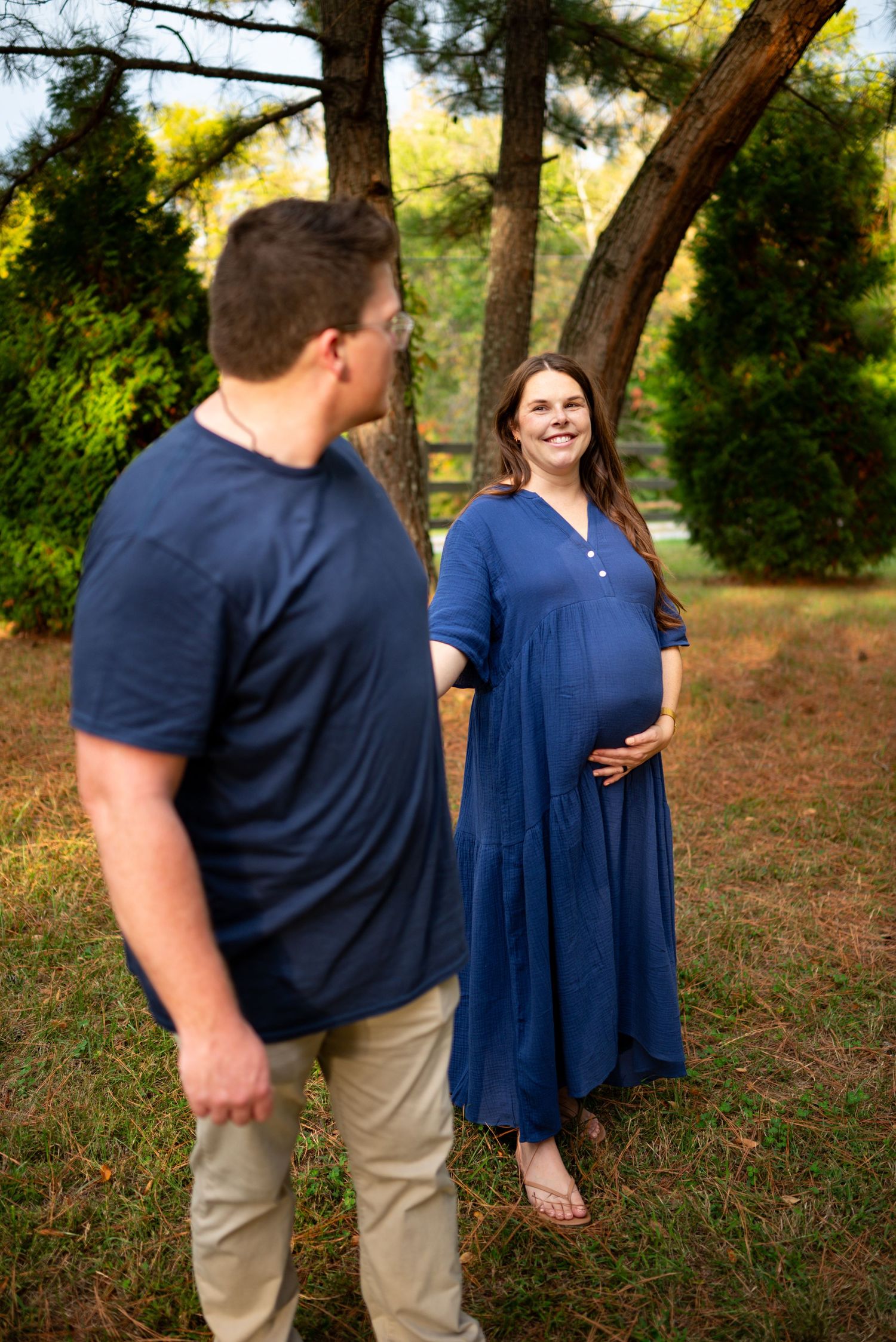 Pregnant woman in blue dress smiles at partner while cradling baby bump in a sunny autumn field.