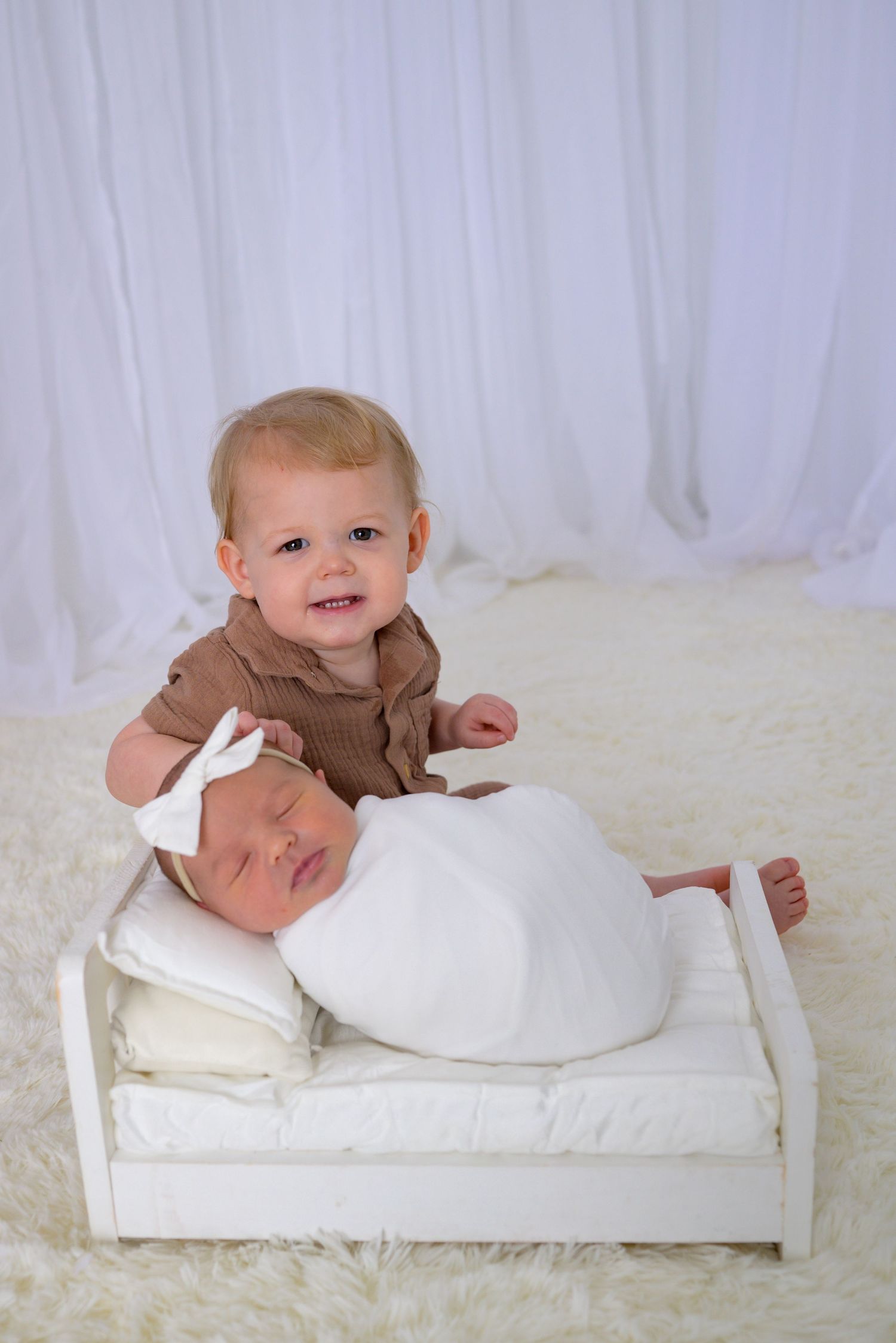 Toddler sibling smiling beside sleeping swaddled newborn baby in a small white bed.