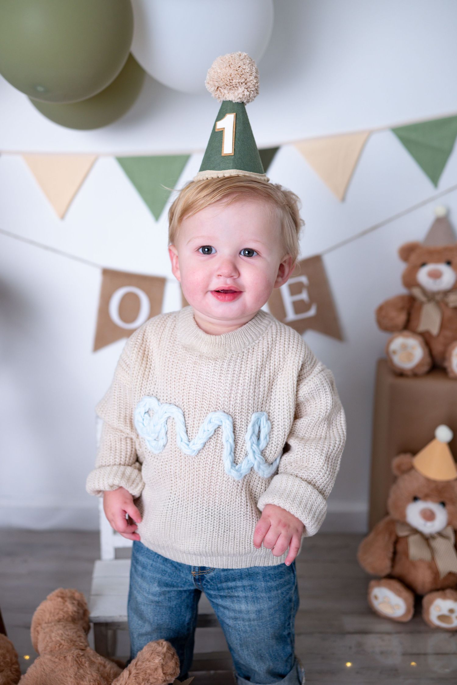Smiling one-year-old boy in cream sweater and party hat stands among teddy bears and green balloon decor.