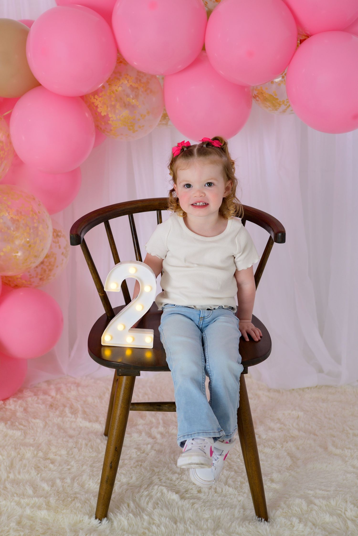A smiling toddler girl sits on a wooden chair holding a lit number 2 sign, surrounded by pink balloons for her 2nd birthday.