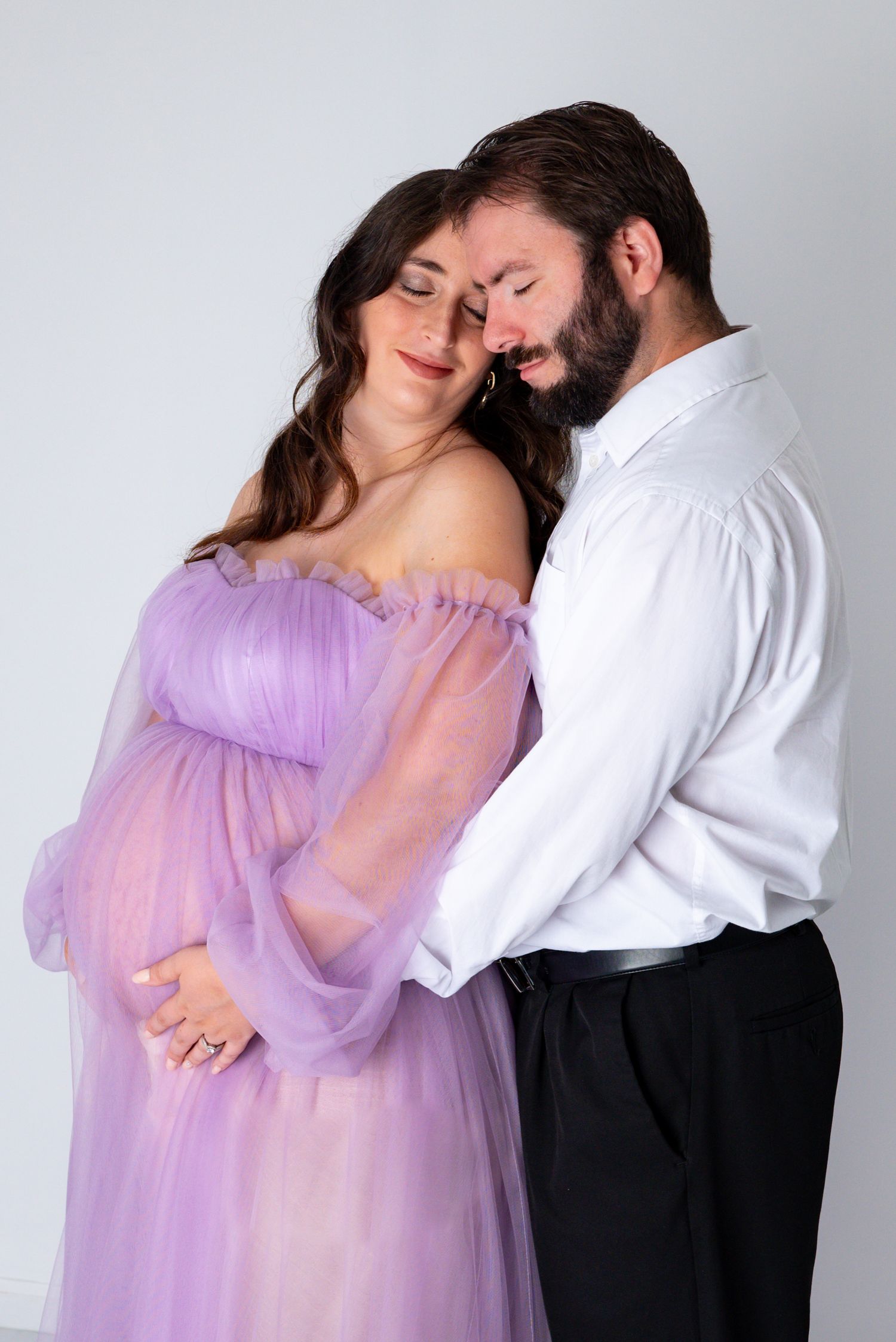A couple shares a tender moment during a maternity session, the woman wearing a flowing lavender tulle gown.