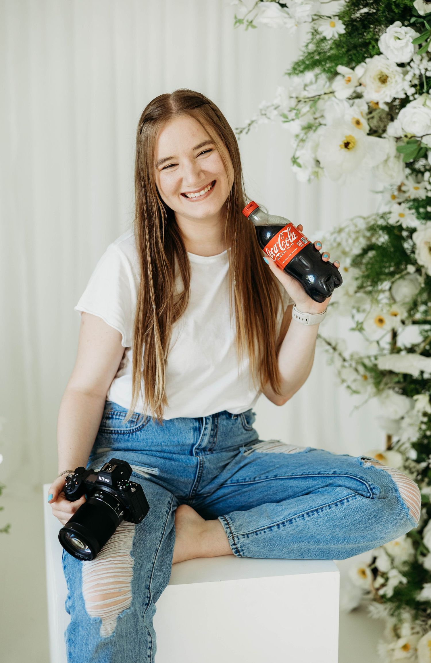 Young female photographer smiling holding Coca-Cola bottle and camera near flowers.