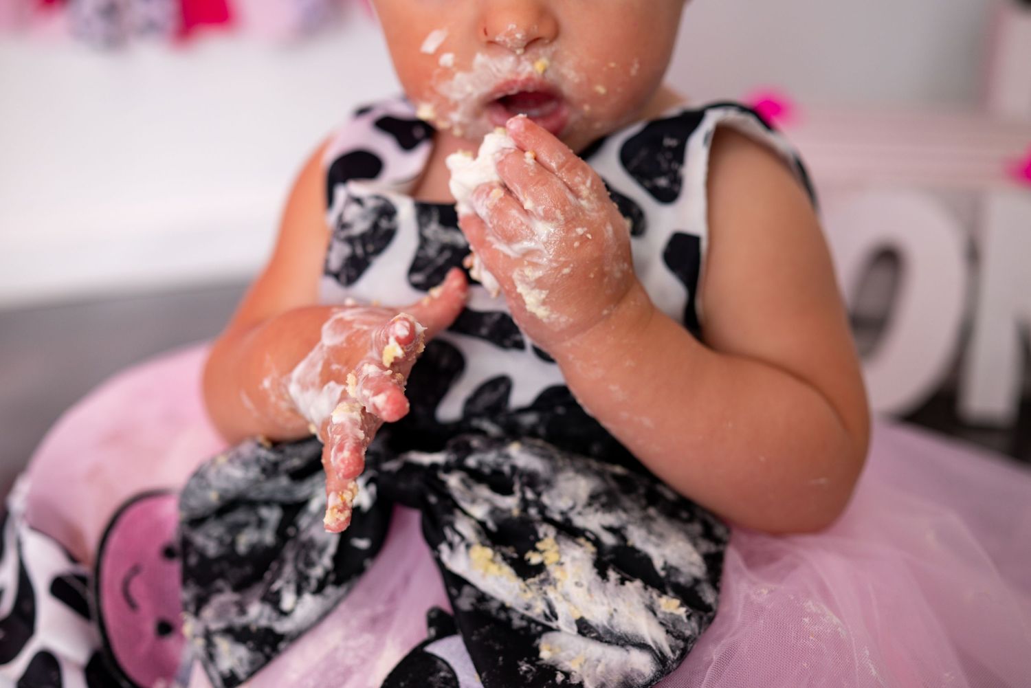 Baby in black white pink outfit enjoys first birthday cake smash with frosting covering hands.