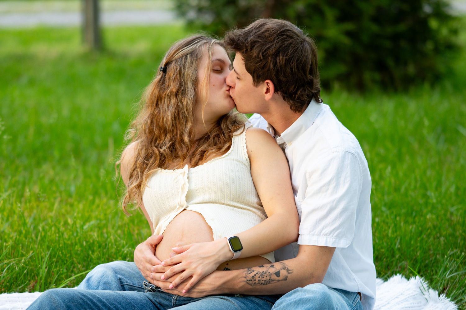 Pregnant couple shares tender kiss while sitting on blanket in lush green park during maternity session.