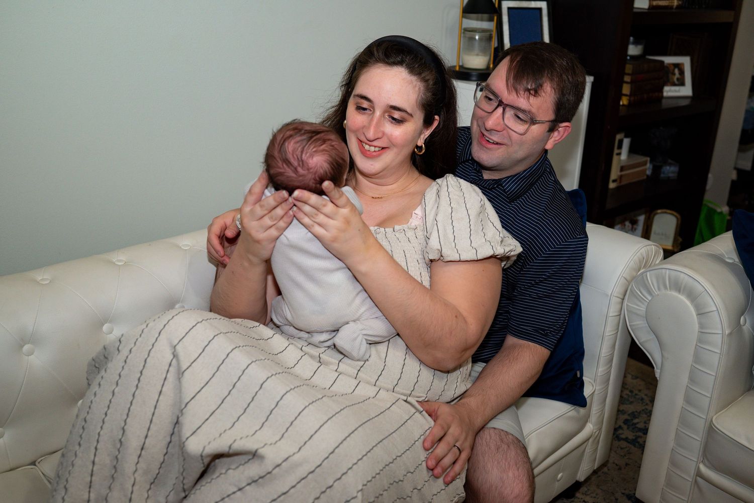 Young couple sitting on white sofa smiling while mother holds newborn baby wrapped in blanket.