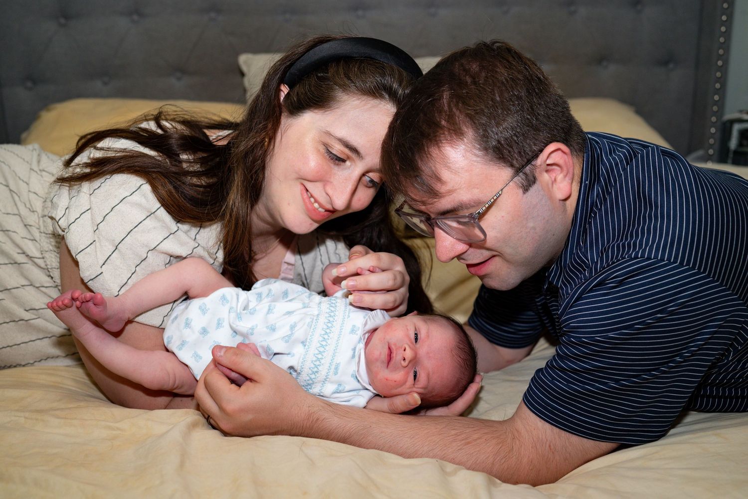 New parents smiling lovingly at their newborn baby lying on a bed in a heartwarming family portrait.