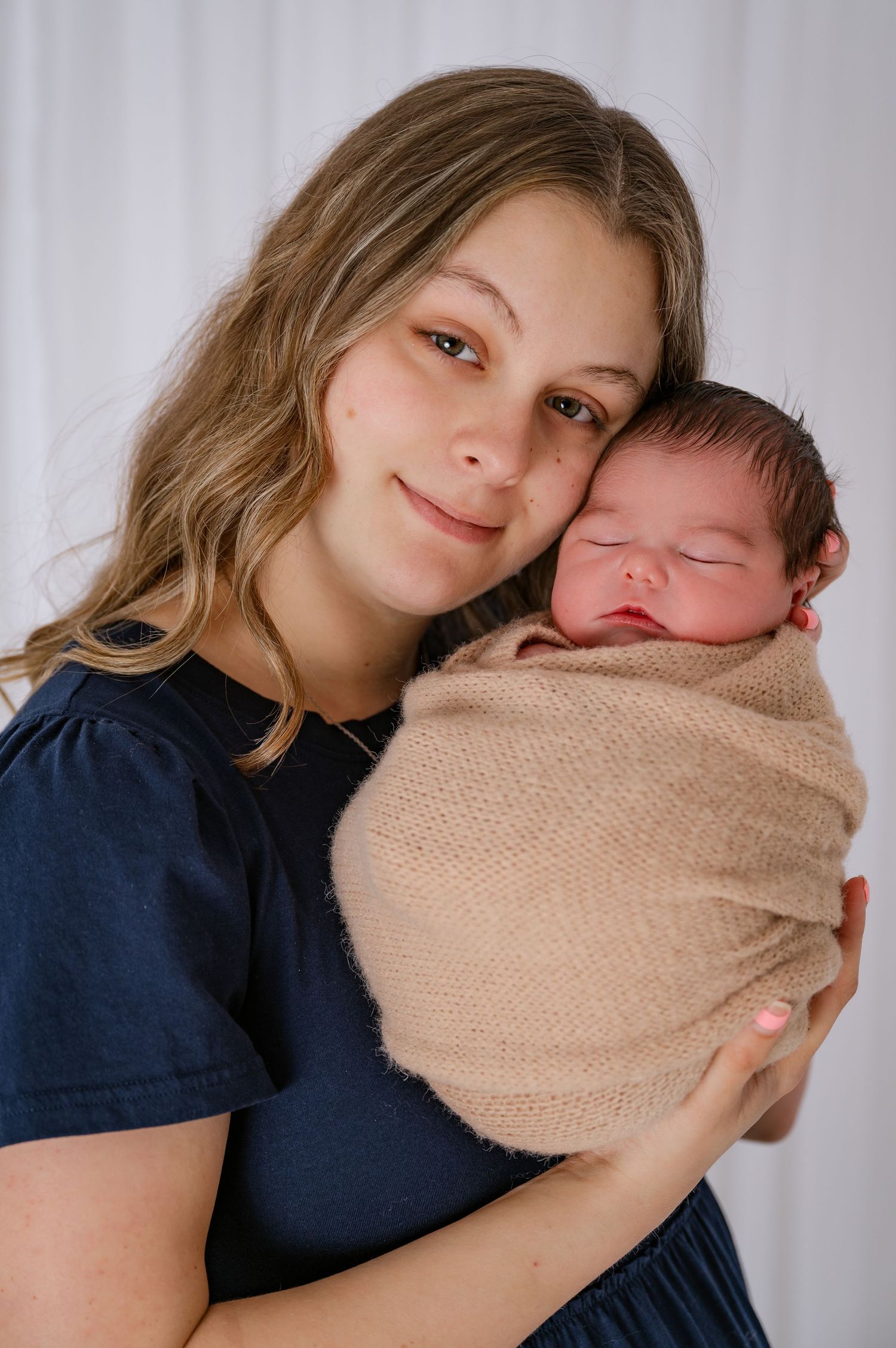 A young mother smiles warmly while cradling her swaddled newborn baby in a beige wrap against a white backdrop.
