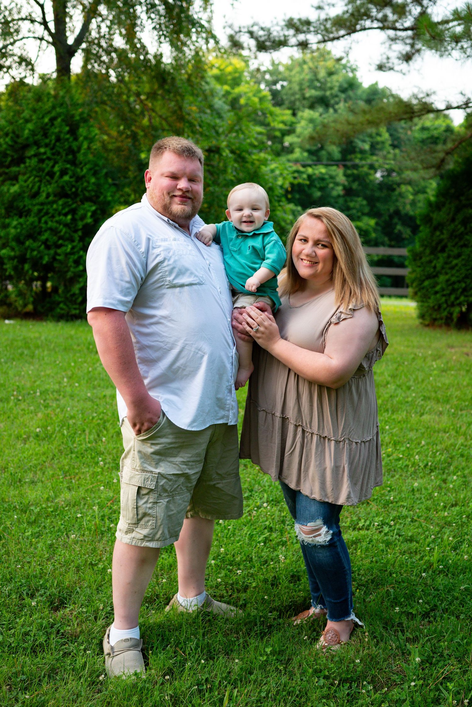 Smiling family of three with baby boy posing together on green grass in backyard.