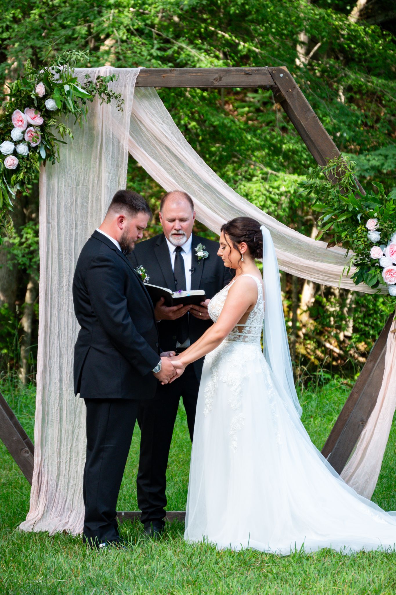 Outdoor wedding ceremony with bride and groom holding hands before officiant under hexagonal arch with floral decor.