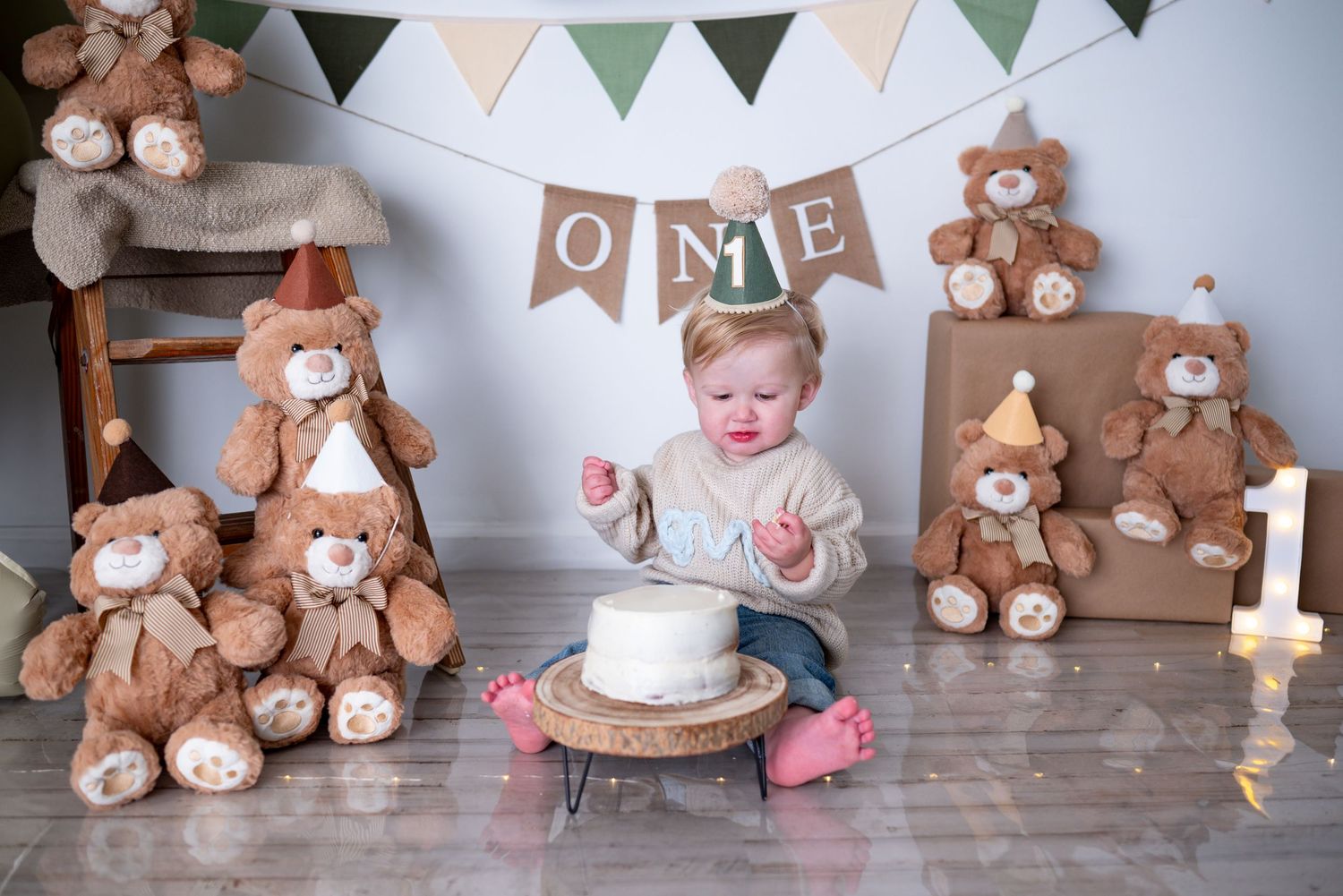 Baby boy in neutral outfit explores first birthday smash cake surrounded by teddy bears and ONE banner.