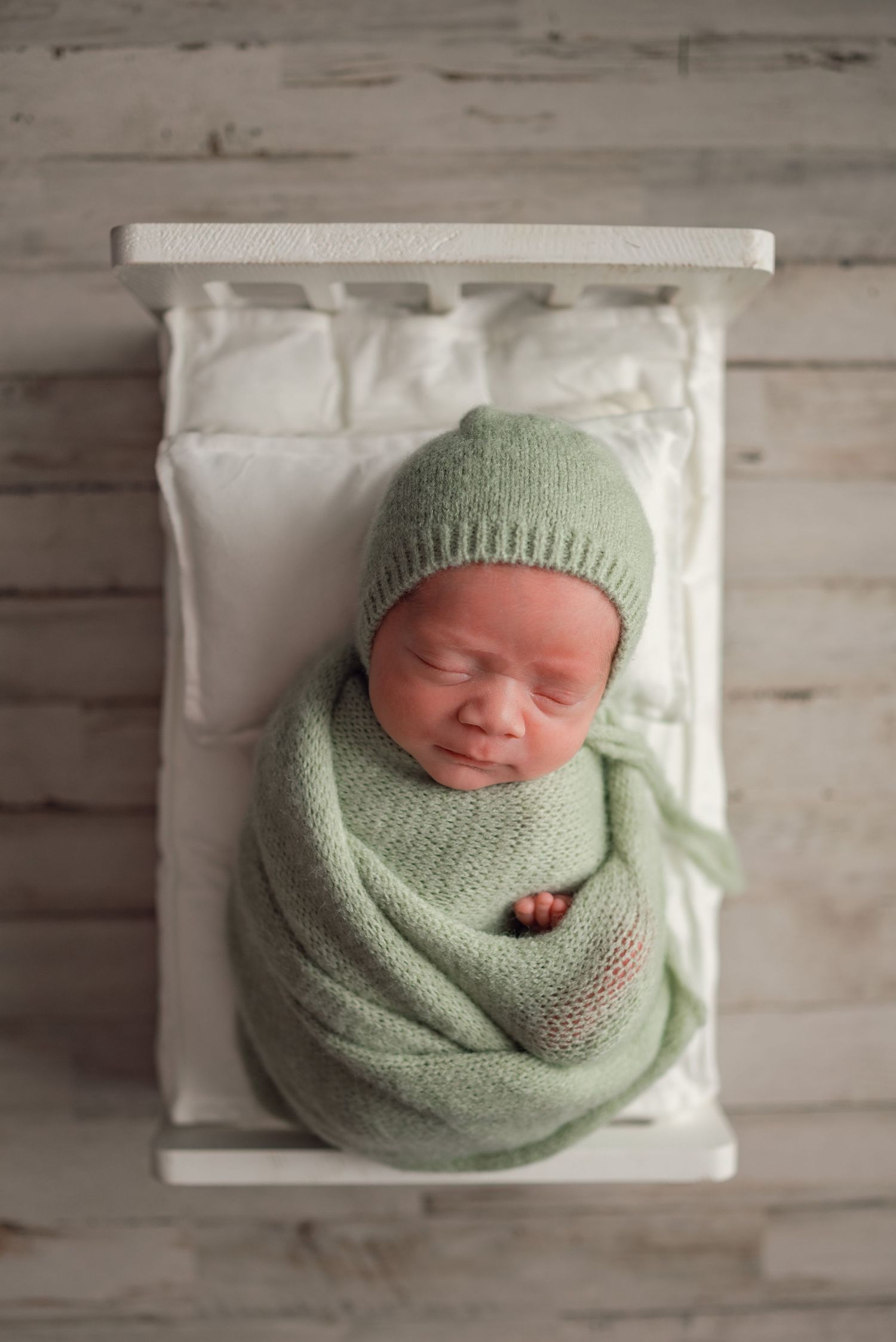Newborn baby swaddled in a soft green knit wrap and matching bonnet, sleeping peacefully in a tiny white bed.