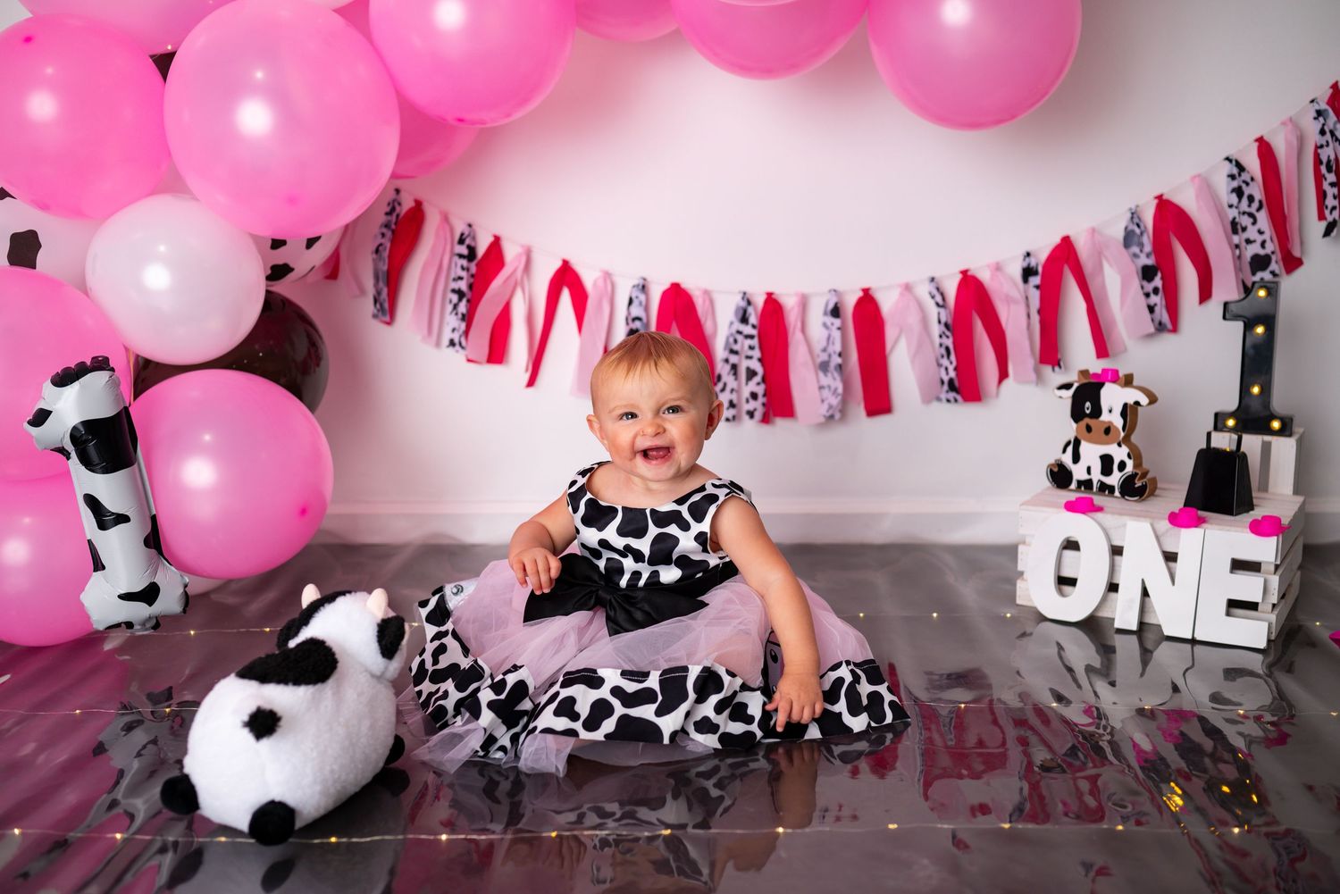 Baby girl in cow print dress sits smiling at first birthday party with pink balloons and ONE sign.