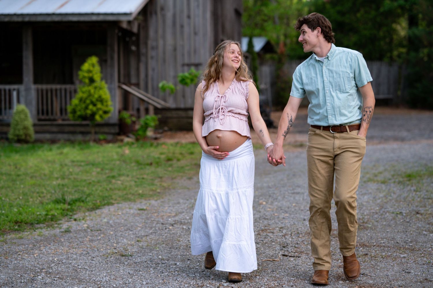 Pregnant woman and her partner walk hand-in-hand along a gravel path near a rustic barn, smiling at each other.