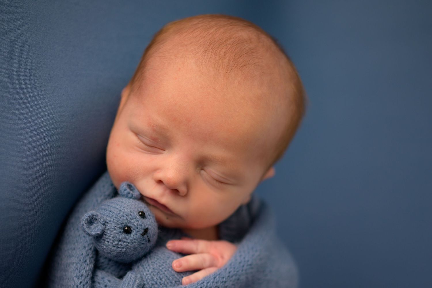 Sleeping newborn baby in blue wrap cuddling a small knitted teddy bear.