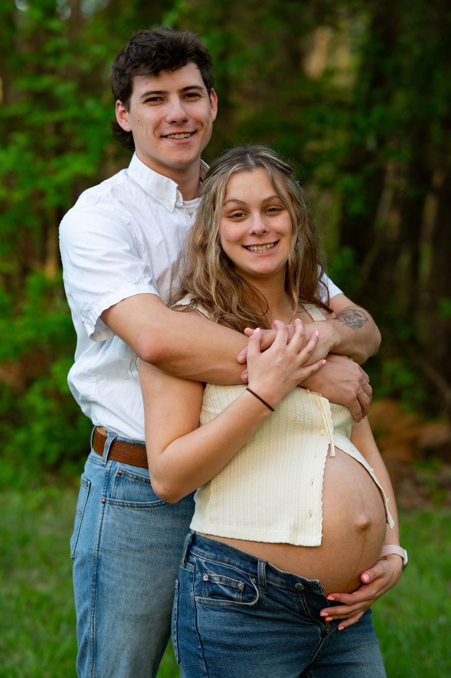 Young couple embracing outdoors during maternity photo session in green wooded setting.