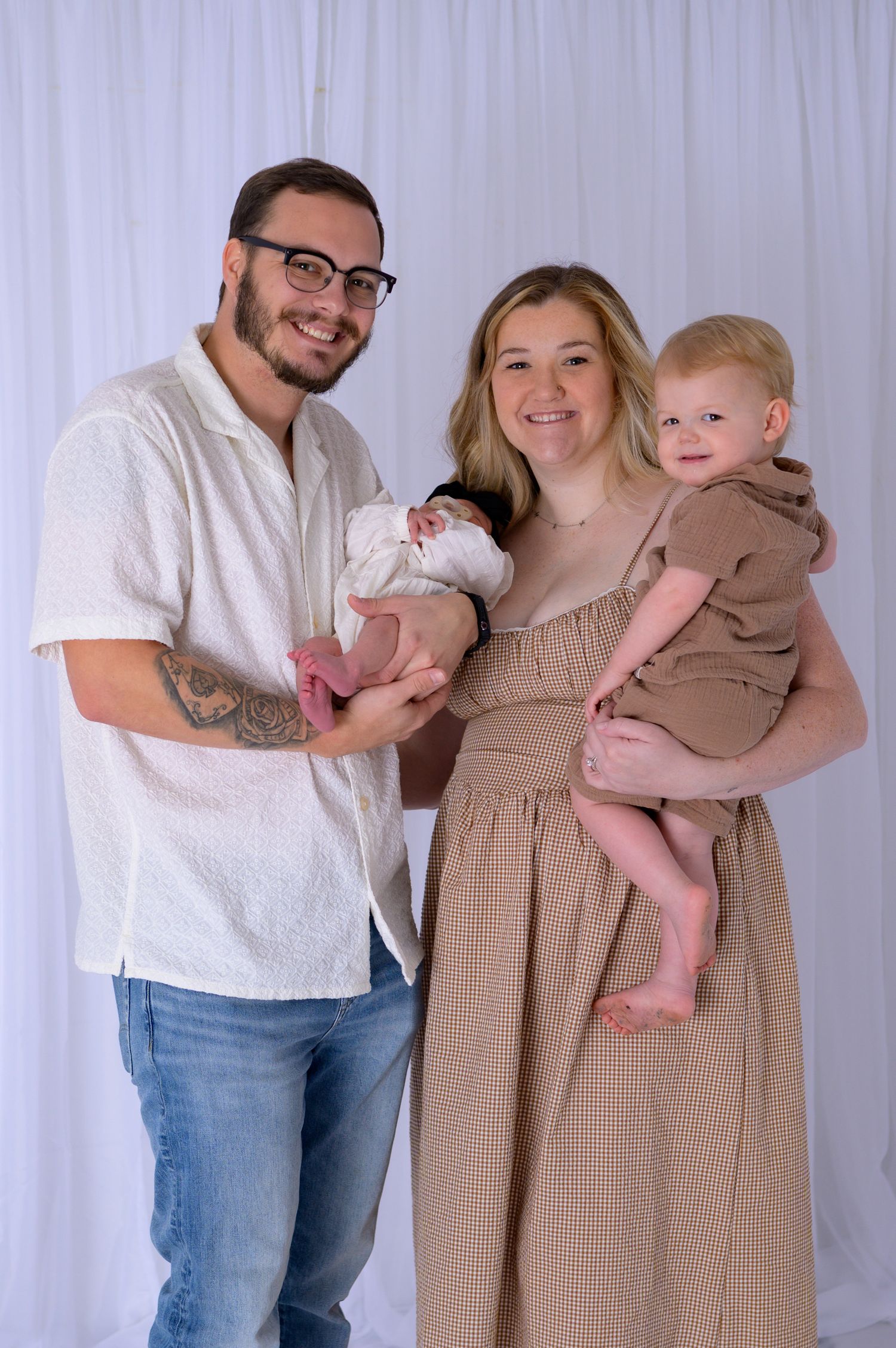 Smiling family of four with newborn and toddler posing against white fabric backdrop.