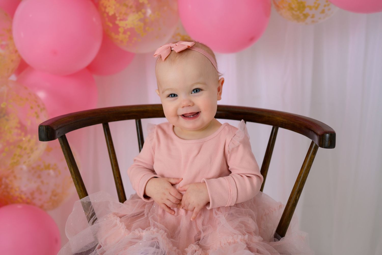 Smiling baby girl in pink dress sitting in wooden chair with pink balloon arch for first birthday.