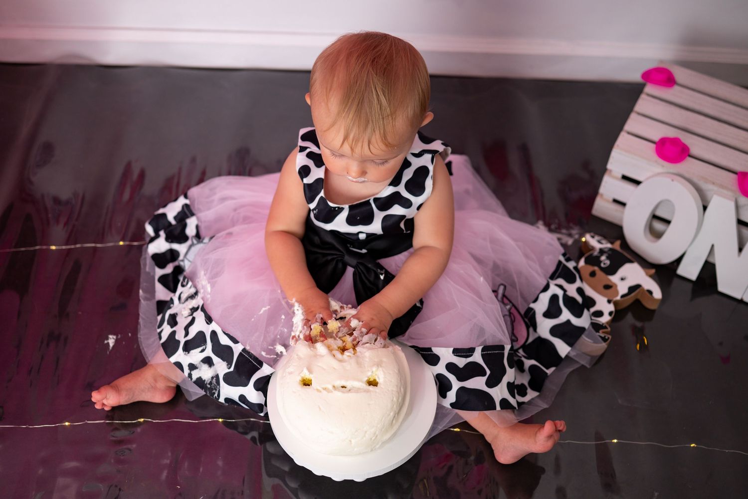Baby girl in cow print tutu smashes white birthday cake during first birthday photoshoot.