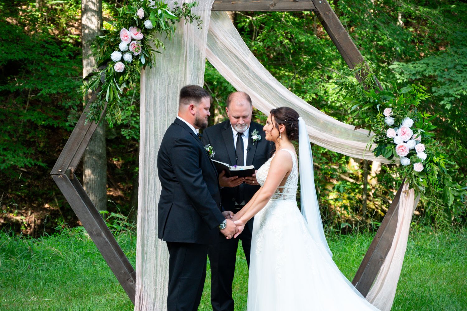Couple exchanging vows outdoors under a rustic wooden arch with white draping and pink floral accents.