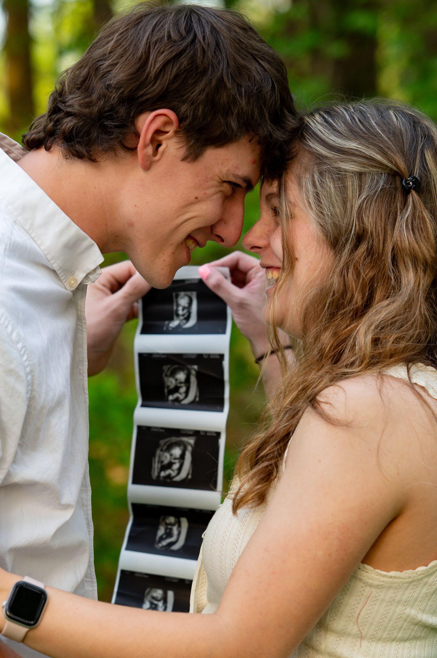 Expecting couple smiling forehead-to-forehead while holding ultrasound photos together in a lush green outdoor setting.