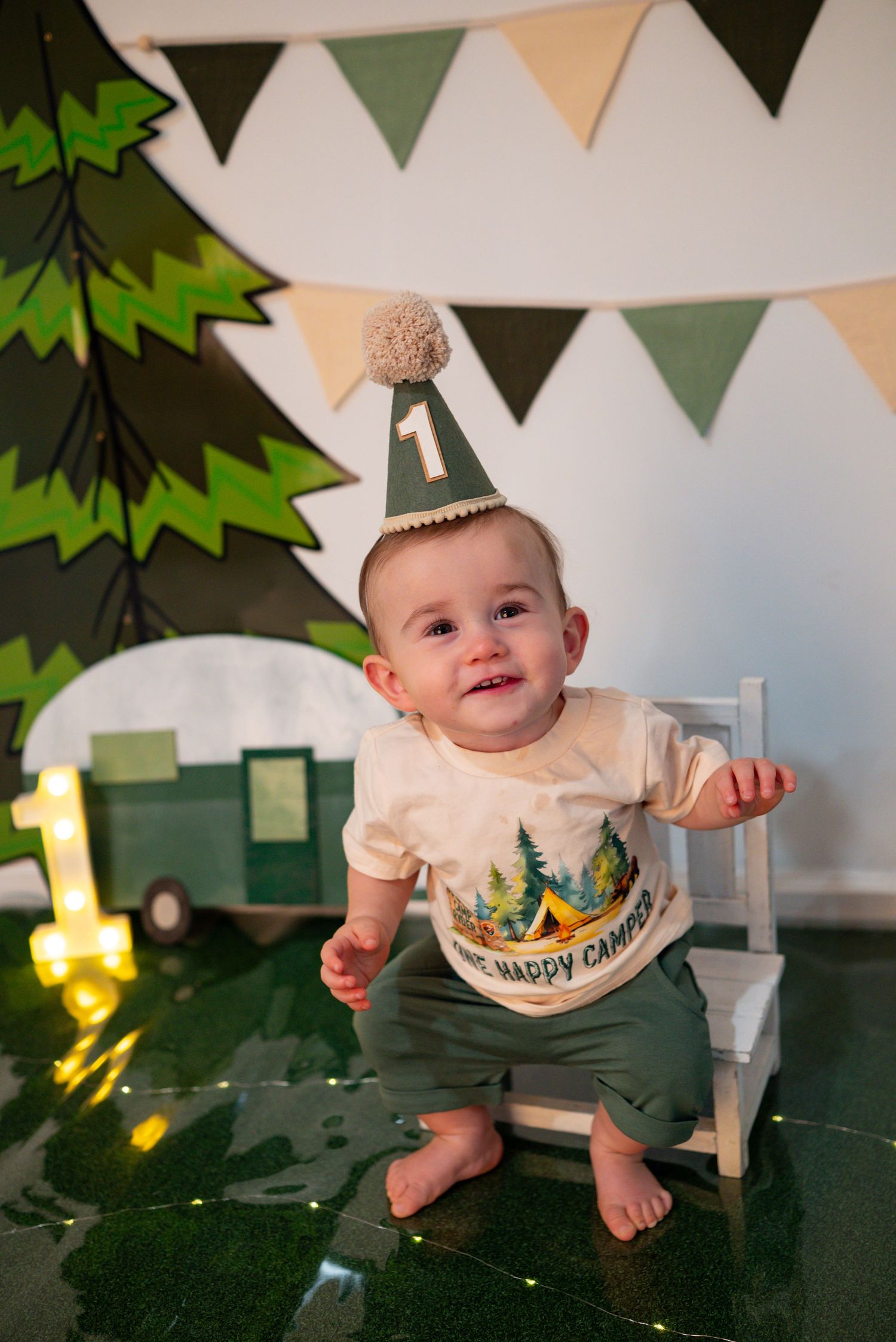 Smiling baby celebrates his first birthday in a camping party hat and Happy Camper shirt with festive decor.
