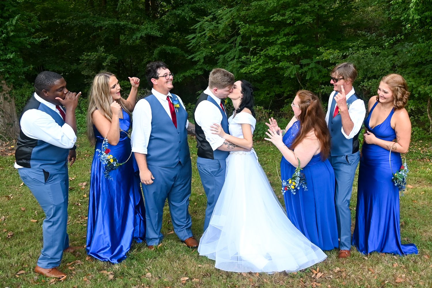 Wedding party in royal blue attire laughing as bride and groom share a kiss outdoors.