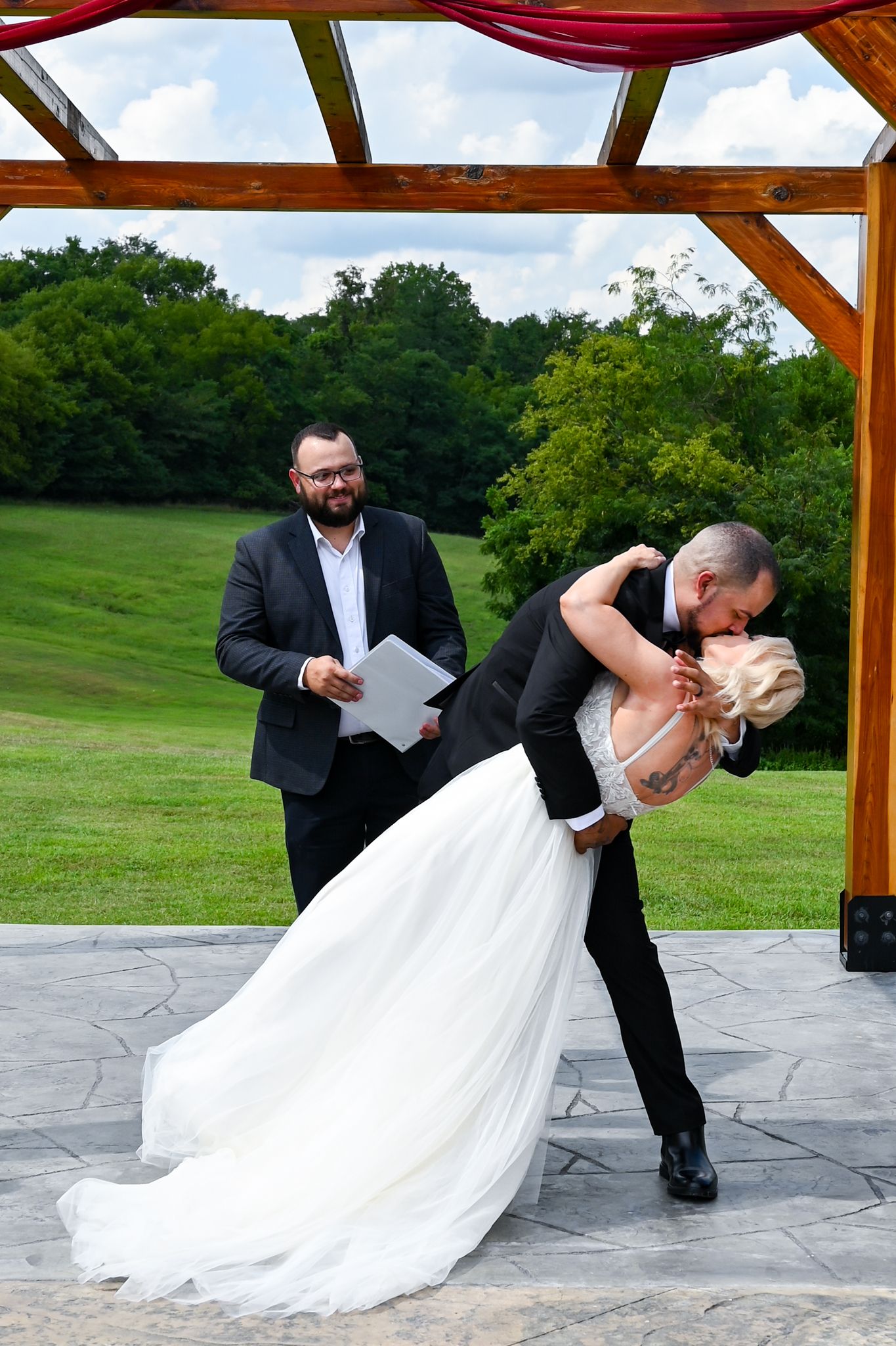 Groom dips bride for a kiss at outdoor wedding ceremony while officiant smiles holding papers at rustic venue.