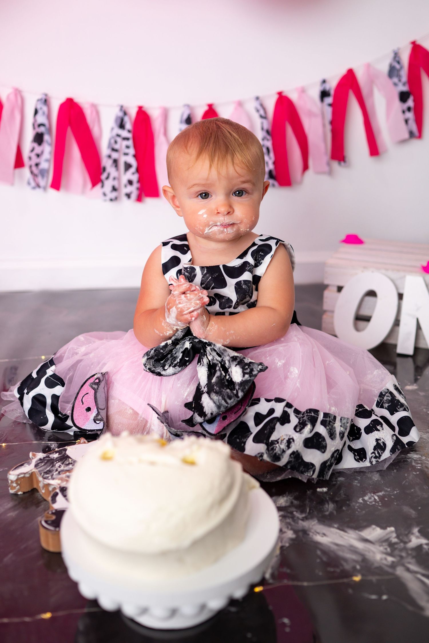Baby girl in cow print tutu dress smashing first birthday cake with frosting on face.