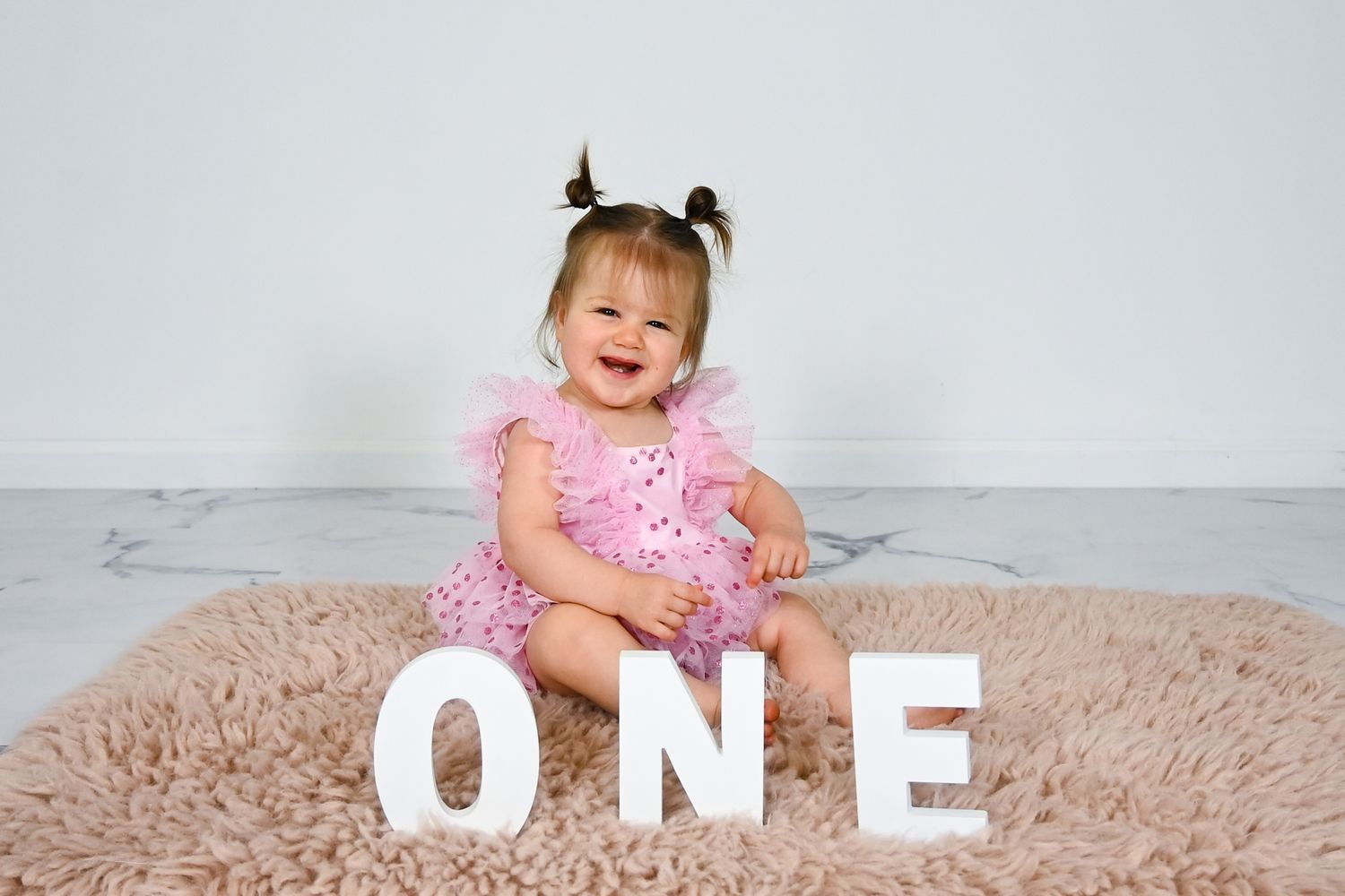 Happy baby girl in pink tutu dress sits on fluffy rug beside white ONE letters for first birthday.