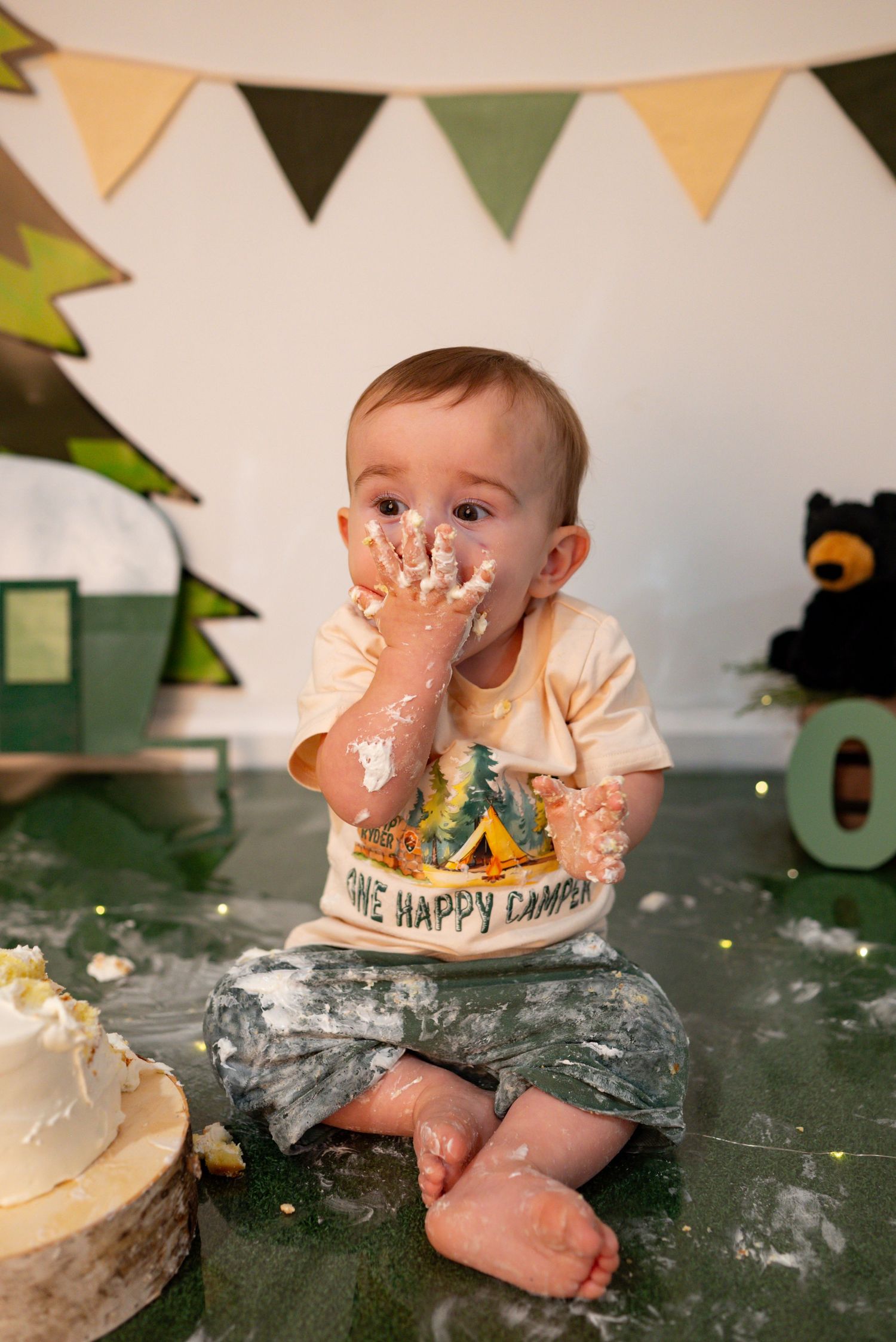 Baby boy smashes his first birthday cake with frosting-covered hands at a camping-themed cake smash session.