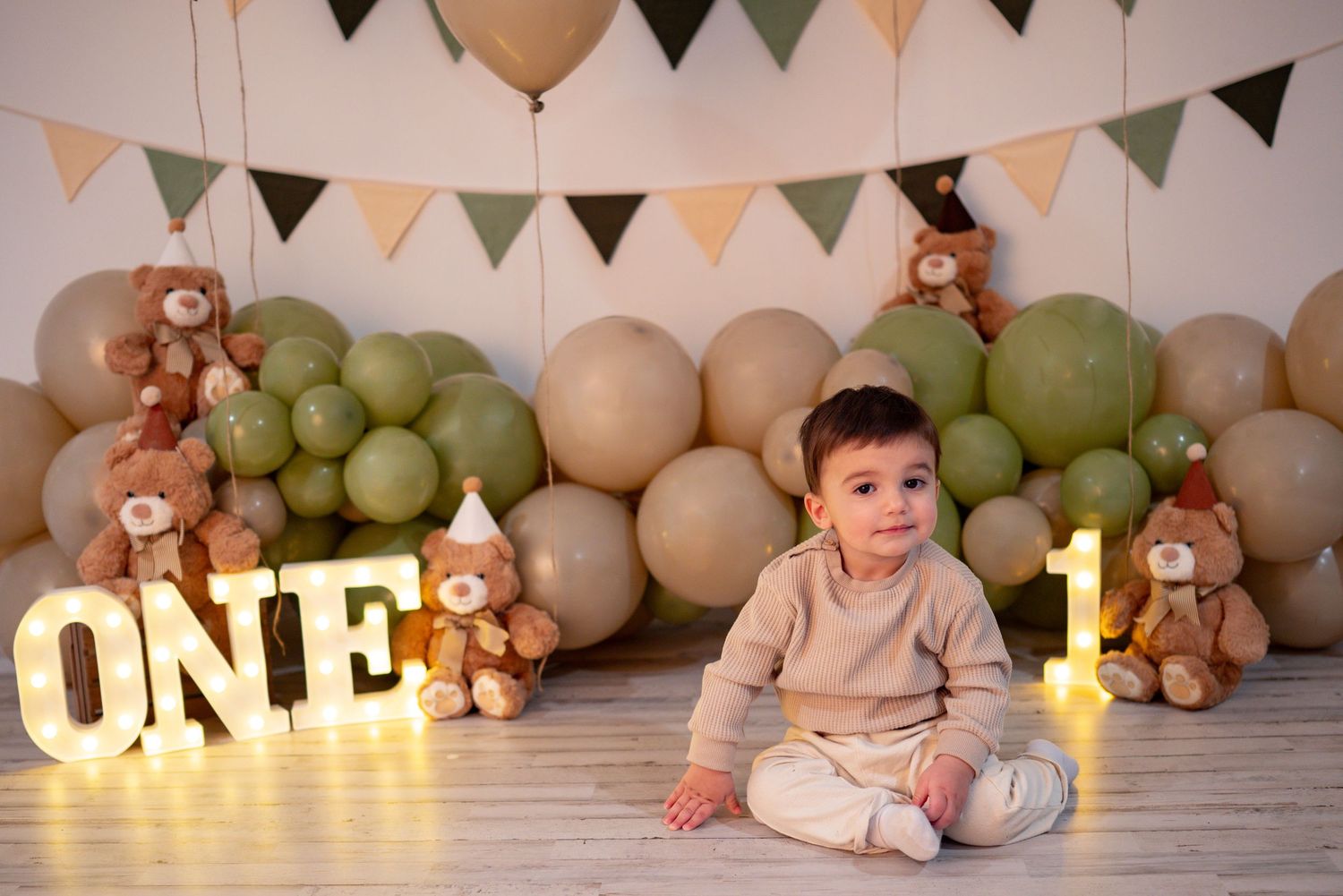 One-year-old boy sits beside ONE marquee letters surrounded by green and beige balloons and teddy bears.