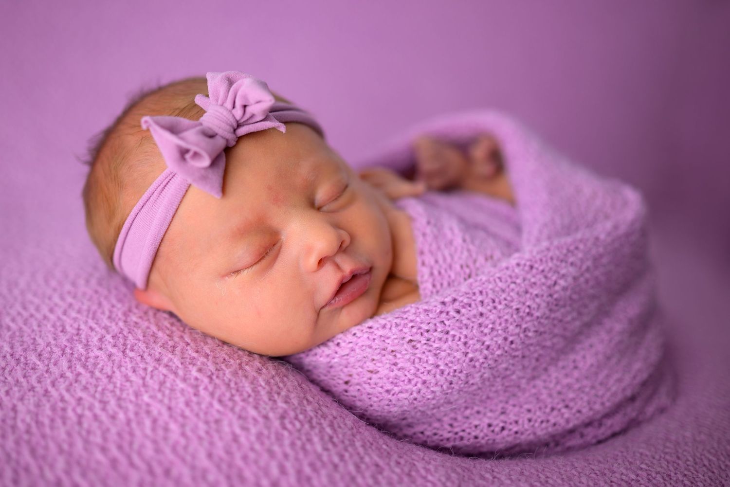 Sleeping newborn baby girl swaddled in lavender knit wrap wearing purple bow headband on purple background.