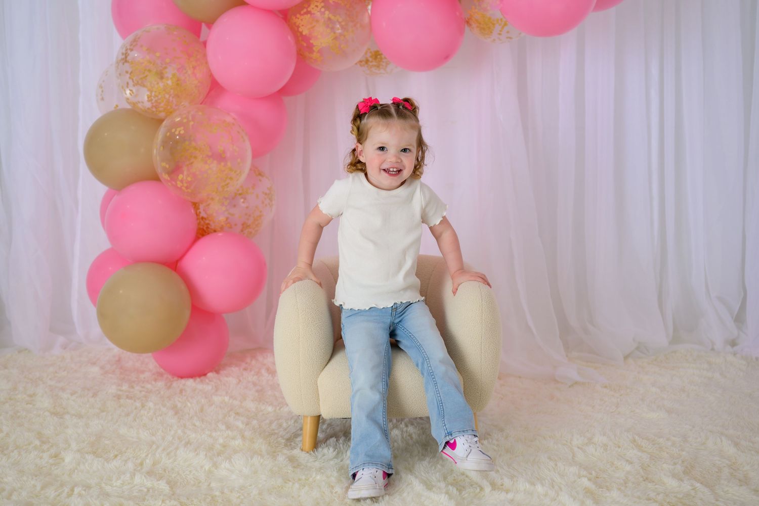 Toddler girl in white tee and jeans sitting on cream chair with pink and gold balloon arch backdrop.