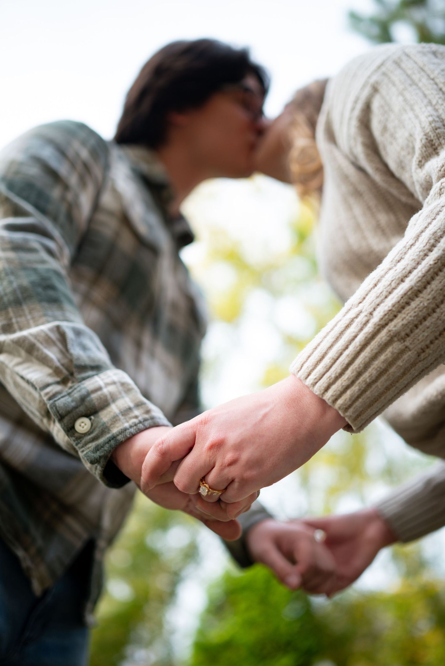 Romantic couple kissing while holding hands outdoors in autumn field, woman wearing gold engagement ring.