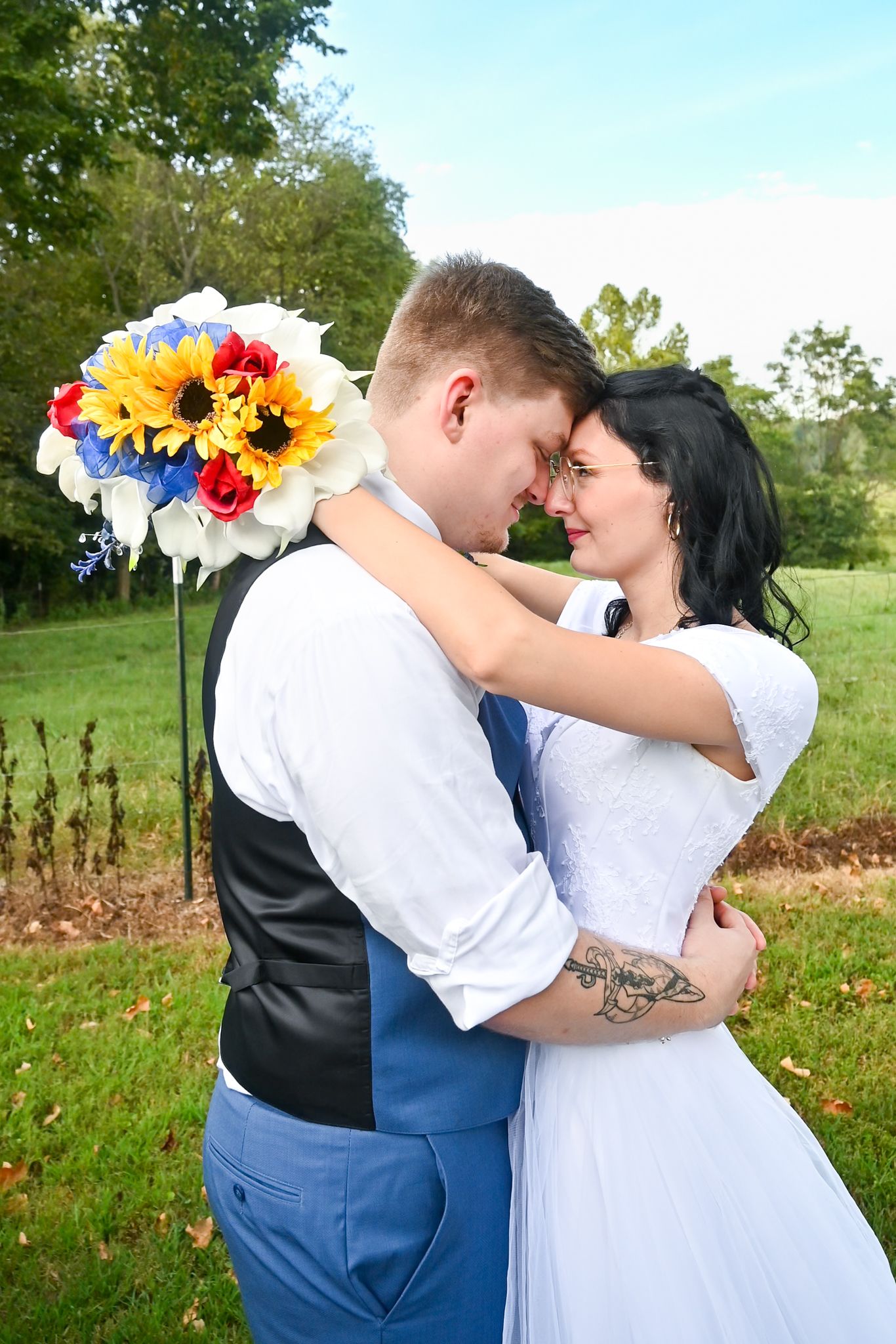 Bride and groom embrace outdoors, touching foreheads romantically, with colorful sunflower bouquet, blue suit, and white wedding dress.