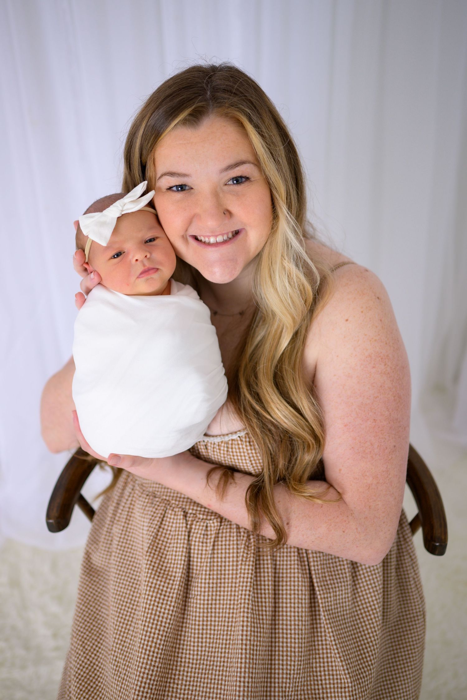 Smiling mother holding swaddled newborn baby girl with white bow headband in professional portrait.