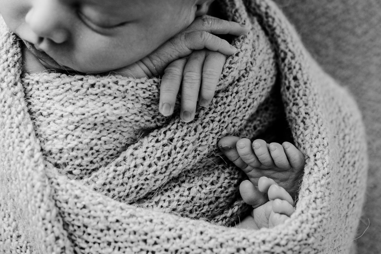 Black and white photo of sleeping newborn swaddled in knit wrap with tiny hands.