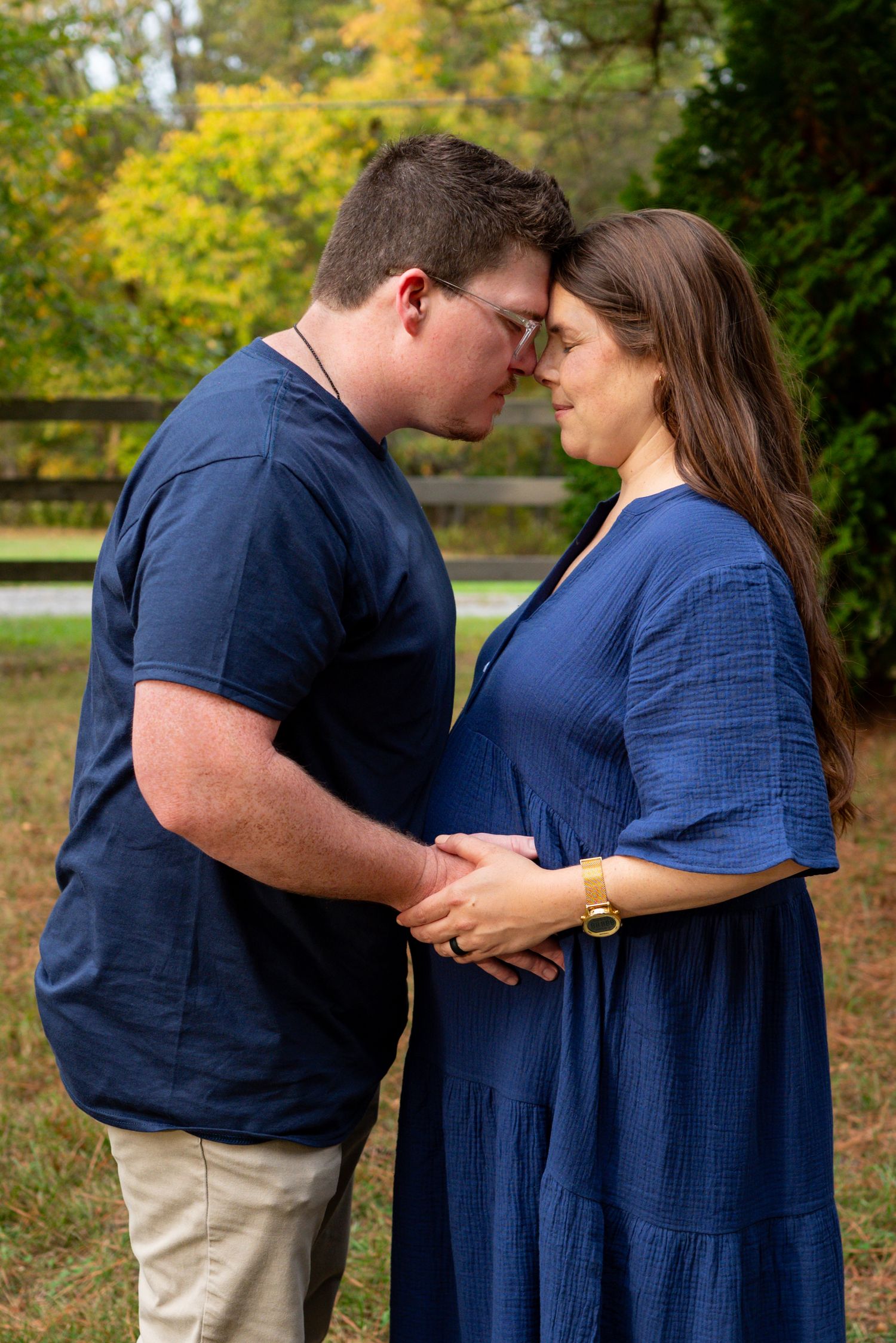 Expecting couple in matching navy blue outfits tenderly touching baby bump forehead to forehead outdoors.