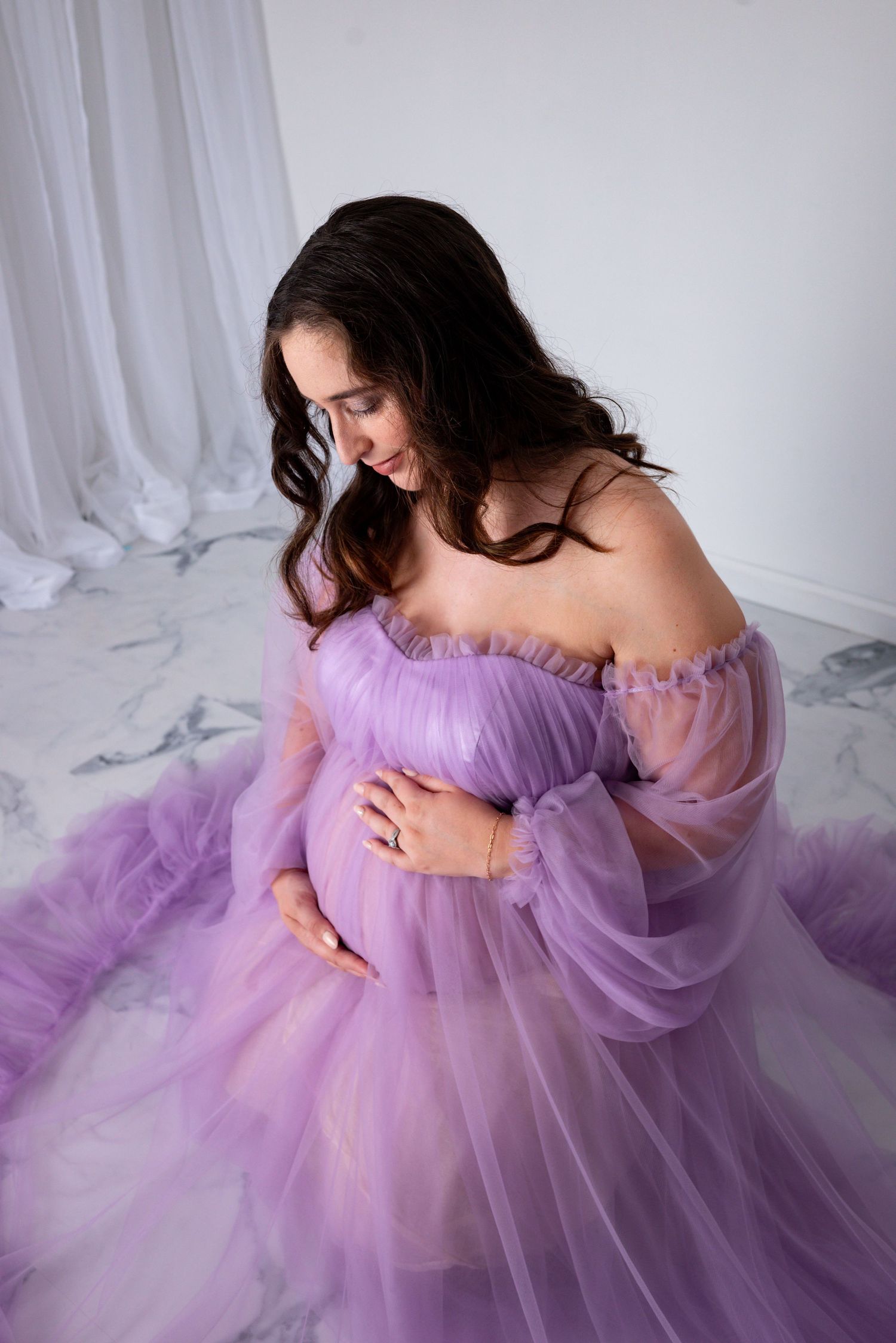 A pregnant woman in a flowing purple tulle gown sits gracefully on a marble floor cradling her baby bump.
