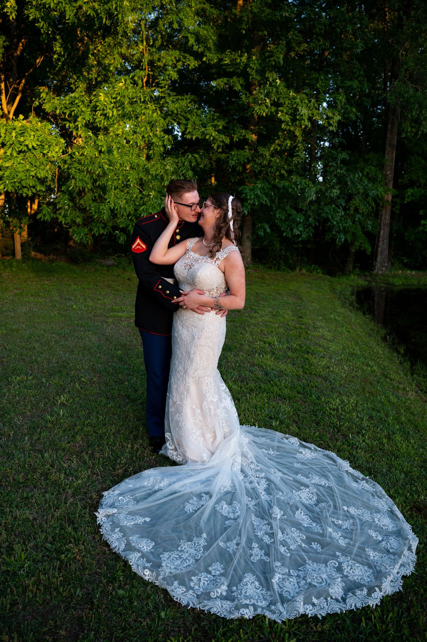 Marine groom in dress uniform kisses bride in lace mermaid gown with cathedral train on green lawn.