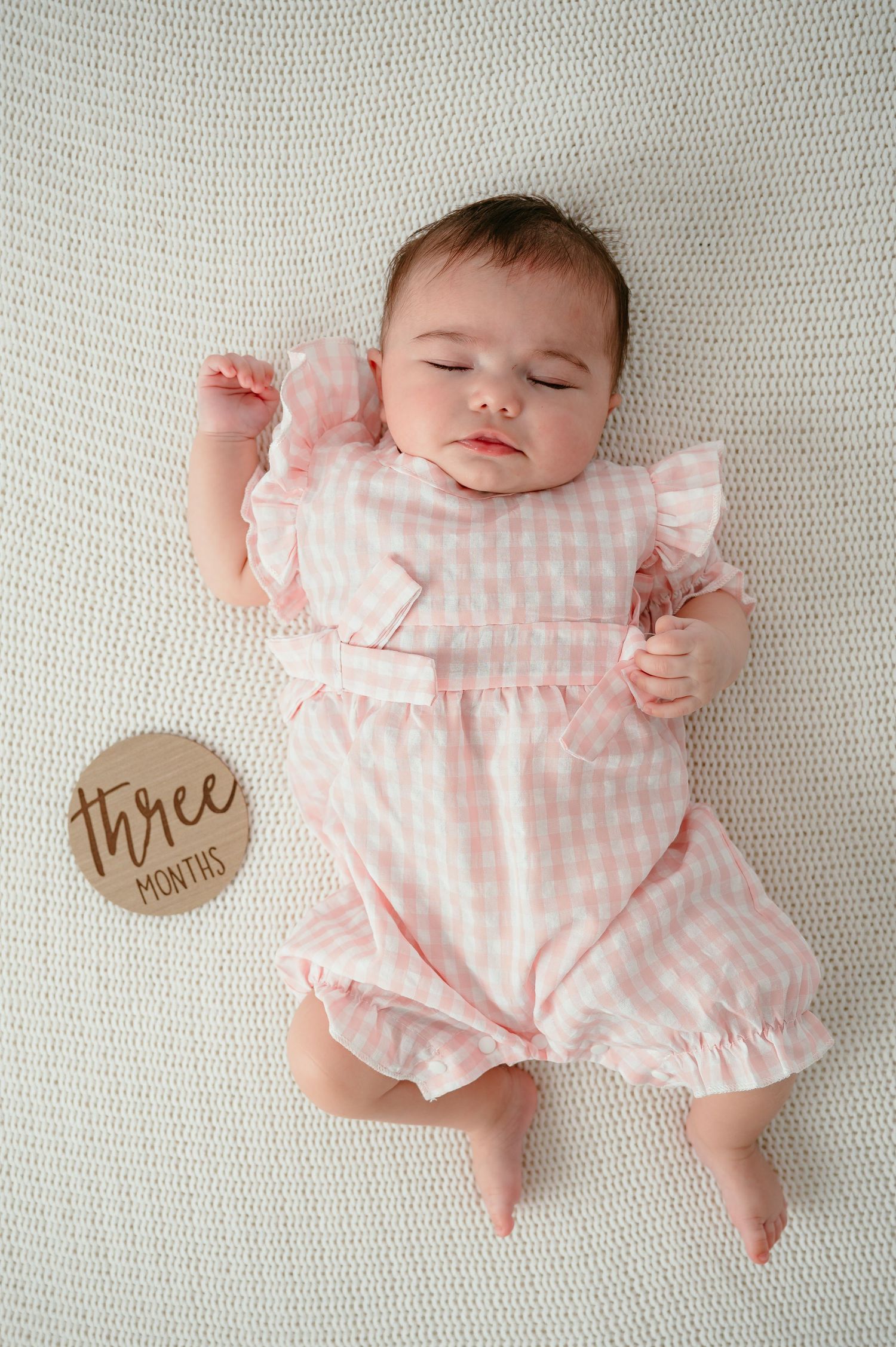 A sleeping three-month-old baby girl in a pink gingham ruffle dress lying on a white knit blanket.
