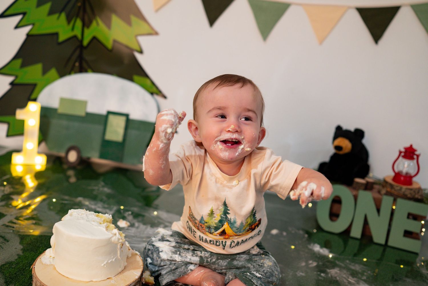 Laughing baby covered in cake frosting celebrates at a camping-themed ONE first birthday cake smash session.