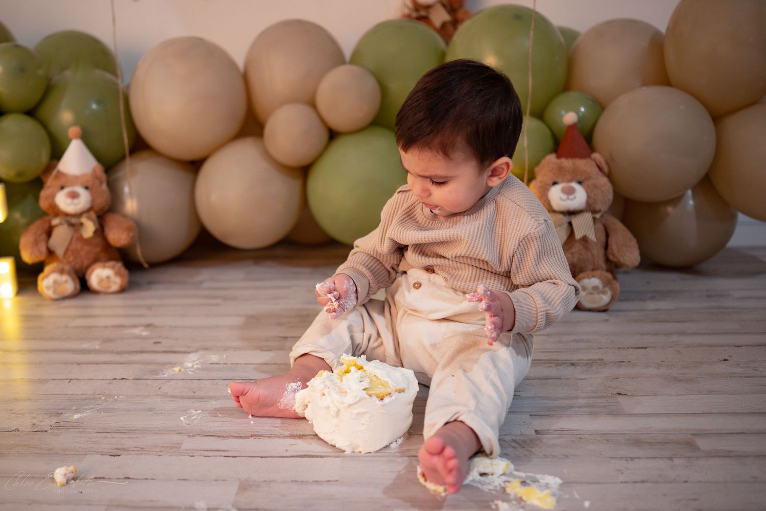 One-year-old boy plays with smashed white birthday cake on the floor in front of green and beige balloons.