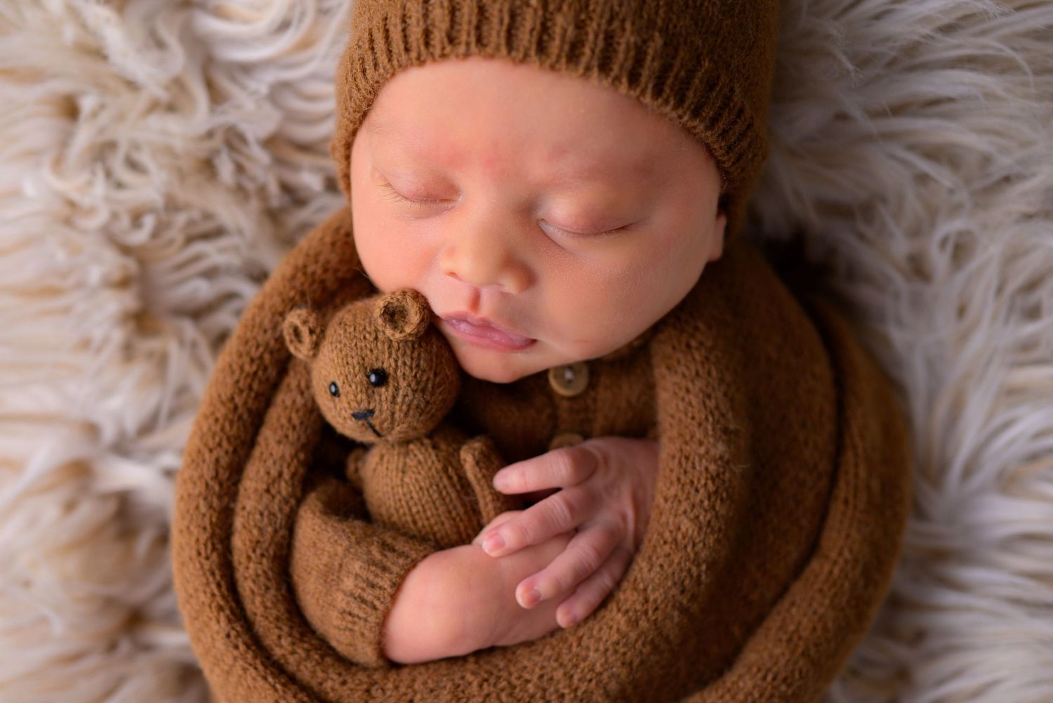Sleeping newborn baby in brown knit wrap and hat cuddling a teddy bear.