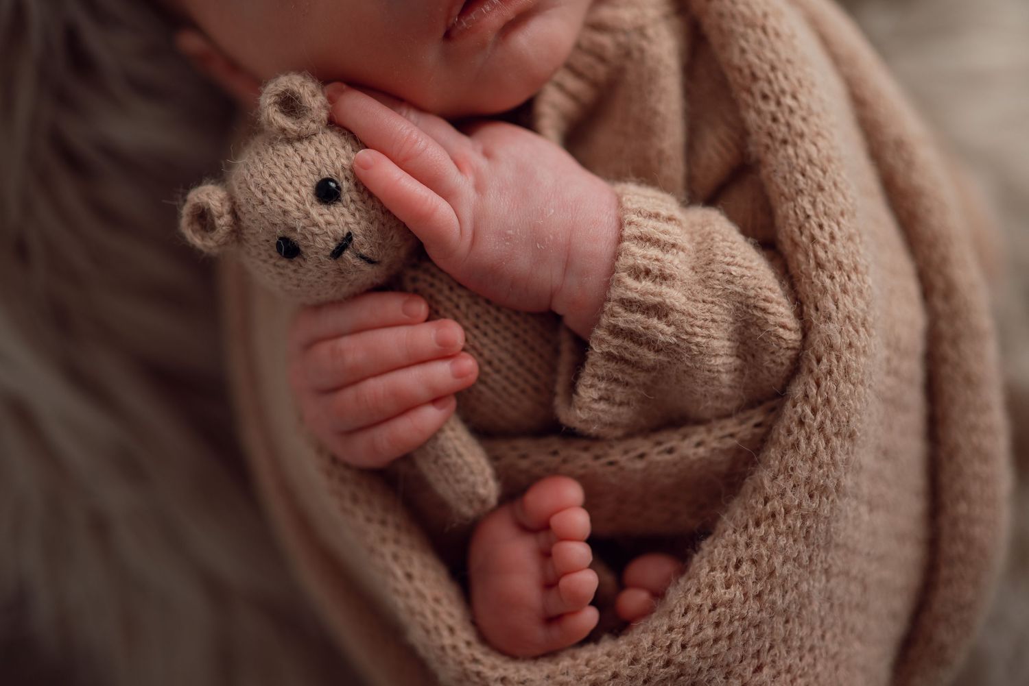 Newborn baby in beige knit outfit holding a small teddy bear, close-up of tiny hands and feet.
