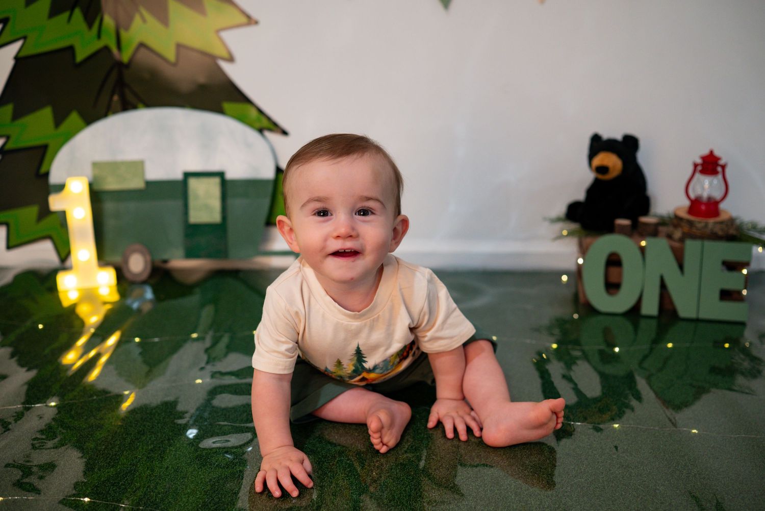 Smiling baby boy lies on green reflective floor with camping-themed first birthday party decorations.