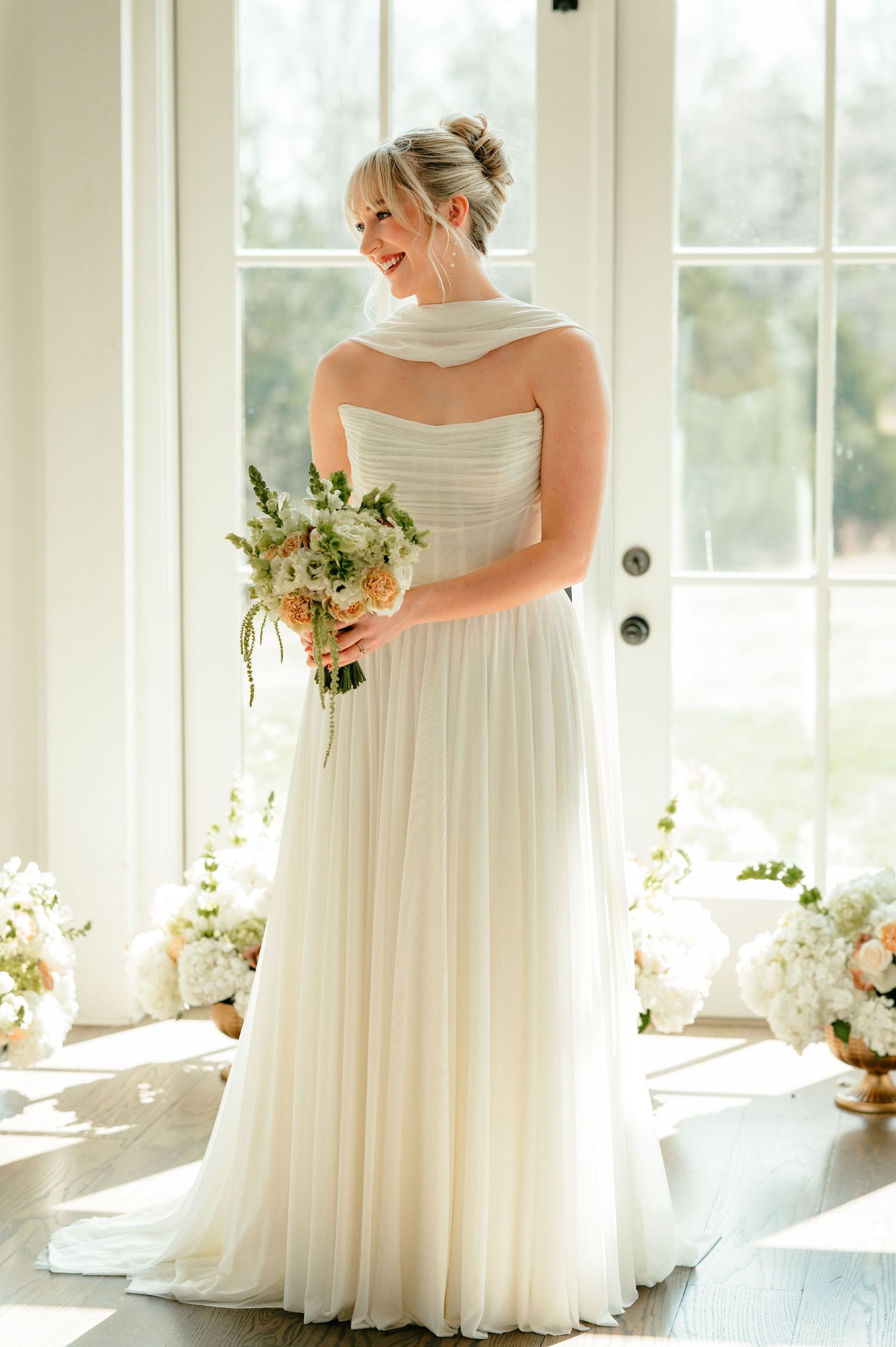 Bride in strapless white chiffon gown holding floral bouquet stands by sunlit French doors in elegant bridal setting.